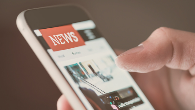 A hand holding a mobile phone displaying news stories.